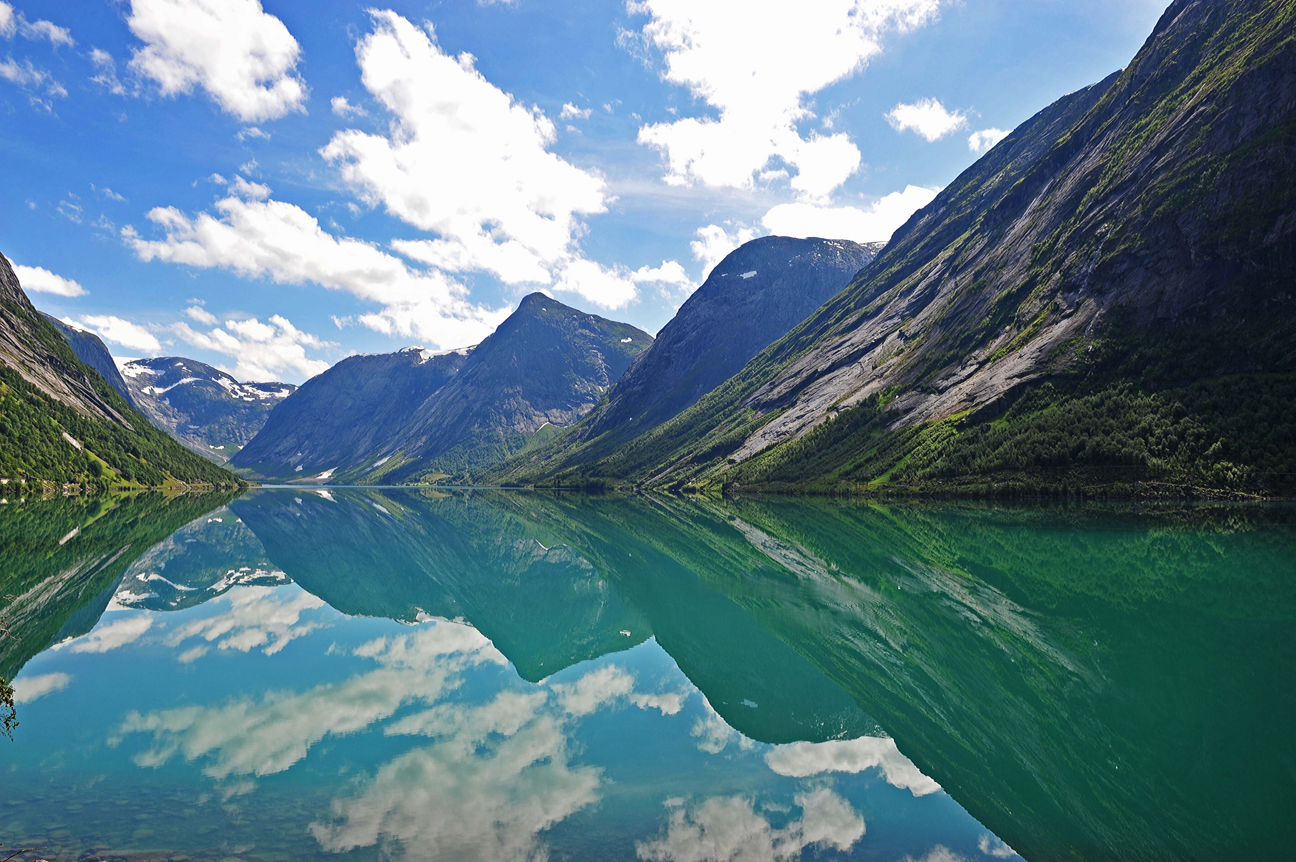 Fjord med sterkr grønn- og blåfarget vann.  Høye fjell på tre sider. Blå himmel med skyer. Landskapet rundt er speilet i vannet,